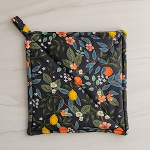 May include: A black quilted hot pad with a floral pattern featuring lemons, oranges, and flowers. The hot pad has a loop handle.