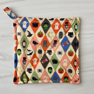 May include: A quilted Halloween-themed pot holder with a diamond pattern. The design features ghosts, pumpkins, bats, and other spooky icons in orange, black, green, and white. It has a small loop for hanging.