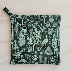 May include: A green pot holder with a white floral and bird design. The pot holder is made of fabric and has a loop for hanging.