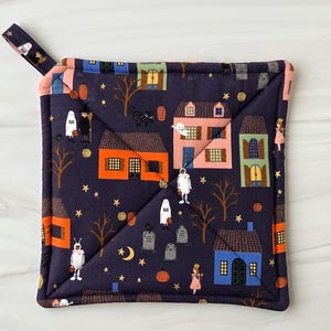 May include: Square navy blue Halloween-themed pot holder with a loop. The fabric features a repeating pattern of houses, ghosts, robots, and trees under a starry sky. The edges are trimmed with a pink border.