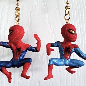 May include: A pair of dangling earrings featuring miniature red and blue Spider-Man figures in dynamic action poses. Each figure is suspended from a gold-toned metal chain and hook, showcasing the character's iconic web-patterned suit and black spider emblem on the chest.
