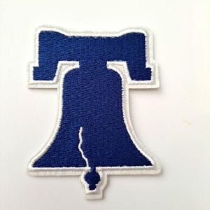 May include: Embroidered patch featuring the Liberty Bell in navy blue with a white border. The bell shape is detailed with a crack and a stylized clapper. This patch is suitable for jackets, bags, or other fabric items.