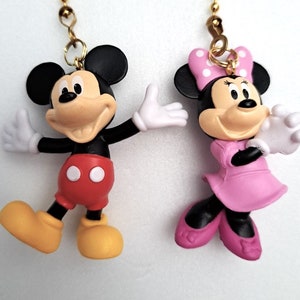 May include: A pair of Mickey Mouse and Minnie Mouse character figurines with gold chains. Mickey Mouse is wearing red shorts with white polka dots and yellow shoes. Minnie Mouse is wearing a pink dress with white polka dots and pink shoes.