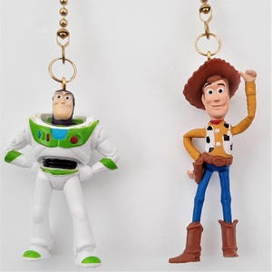 May include: Two ceiling fan pull chains featuring miniature plastic figures of Buzz Lightyear and Woody. Buzz is in a white, green, and purple space suit. Woody wears a brown cowboy hat, yellow shirt with cow print vest, and blue pants.