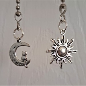 Peut inclure: Deux chaînettes de tirage de ventilateur argentées avec des breloques décoratives. Un breloque est un croissant de lune avec un chat assis dessus. L'autre breloque est un soleil avec des rayons.