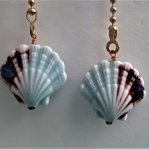 May include: Two seashell-shaped ceiling fan pulls. The ceramic shells are light blue with dark brown and blue accents. Each shell is attached to a gold-colored chain with a decorative connector.