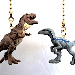May include: Two dinosaur-themed fan pulls. One is a brown Tyrannosaurus Rex with an open mouth, the other is a gray Velociraptor with blue stripes. Both are attached to gold-colored chains.