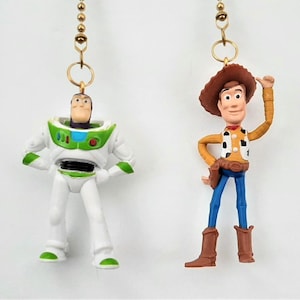 May include: Two plastic figurines of characters from the Toy Story movie franchise. The figurine on the left is Buzz Lightyear, a space ranger, wearing a white and green spacesuit. The figurine on the right is Woody, a cowboy, wearing a brown hat, a yellow vest, and blue jeans.