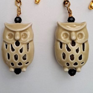 May include: A pair of cream-colored ceramic owl earrings with black bead accents. The owls have a cut-out design and are hanging from gold-colored chains.