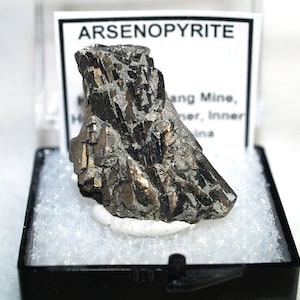May include: A metallic, silver-grey arsenopyrite mineral specimen with sharp, angular crystalline structures. The mineral is displayed inside a black plastic box with a white label reading ARSENOPYRITE and additional location text.