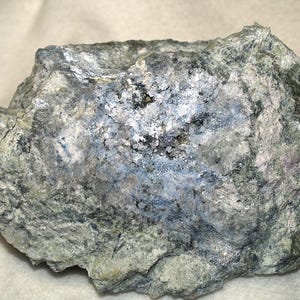 May include: A roughly shaped rock sample displaying a blend of grey, blue, and white tones. The surface has a textured look with visible mineral deposits. The rock's irregular form and varied colours suggest a natural, unpolished state.
