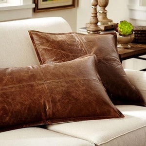 May include: Three brown leather throw pillows with stitched details rest on a cream-colored sofa. The pillows have a distressed, textured appearance, suggesting a vintage or rustic aesthetic. The pillows are rectangular and square.