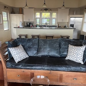 May include: A dark blue leather couch with a wooden frame and drawers. The couch has three cushions and two throw pillows with a geometric pattern. The couch is in front of a kitchen with white cabinets and a stainless steel refrigerator.