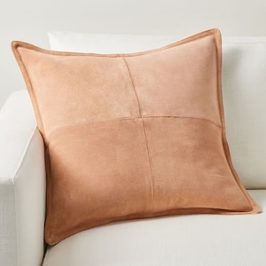 May include: A light brown suede pillow with a square stitched design. The pillow is on a white couch.