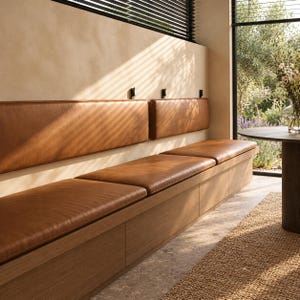 May include: A long, built-in bench featuring a light brown wooden base and smooth, tan leather seat cushions. The bench includes matching tan leather back cushions mounted to the wall with small black hardware, creating a continuous seating area.