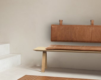 May include: A wooden bench with a brown leather cushion and backrest. The bench is in front of a white wall and has a light brown wood floor.