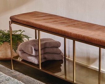 May include: A gold metal bench with a brown leather seat and a lower shelf. The bench has a woven basket with a green plant on the left side and a stack of folded blankets on the right side.