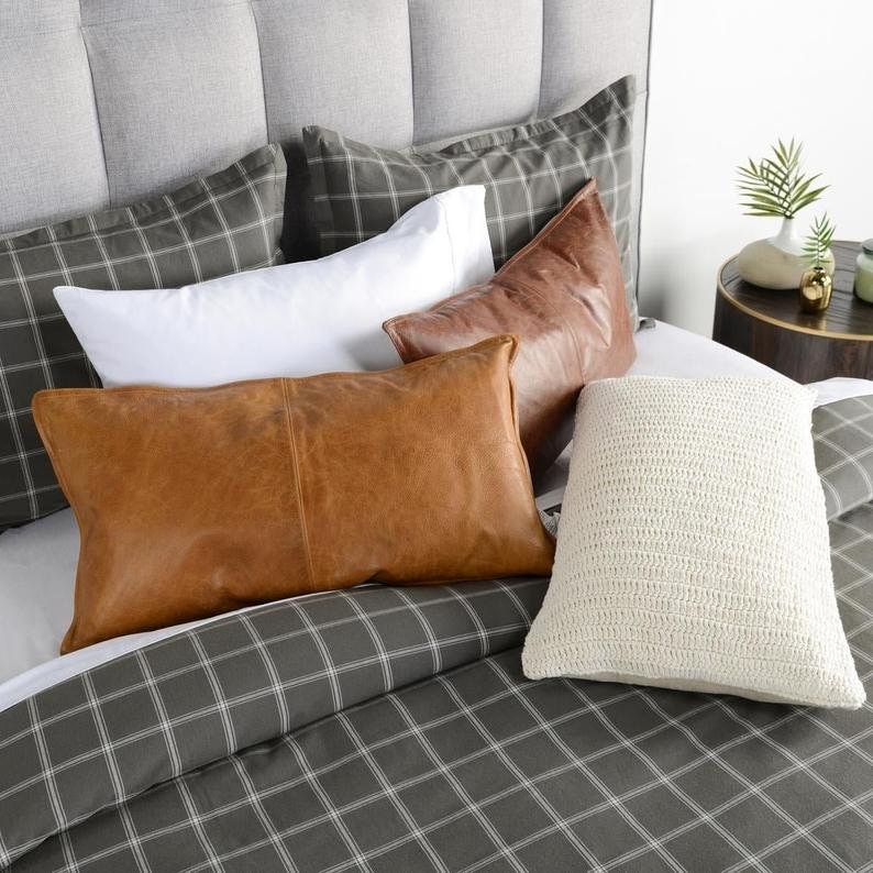 May include: A bed with a gray upholstered headboard and a gray plaid comforter. The bed is decorated with several pillows, including white, brown leather, and cream-colored crochet pillows.