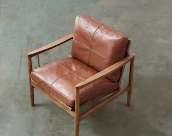 Op de afbeelding: Een bruine leren fauteuil met een houten frame. De stoel heeft een eenvoudig, modern ontwerp met een comfortabele, gepolsterde zitting en rugleuning.