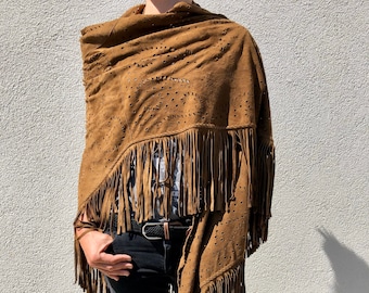 May include: A brown suede shawl with fringe and decorative cut-out patterns. The shawl is draped over the shoulders. The person is wearing black jeans and a black belt.