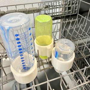 Może przedstawiać: Trzy butelki do karmienia niemowląt z białymi plastikowymi uchwytami znajdują się w zmywarce. Butelki są wykonane z przezroczystego szkła z niebieskimi i białymi oznaczeniami. Jedna butelka jest zielona.