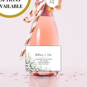 Puede incluir: Una pequeña botella de champán rosado con una pajita a rayas doradas y blancas, una cinta dorada y una etiqueta blanca con el texto "Bethany & Jack Felicidades por su compromiso 20.10.2022".