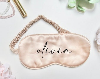 May include: A light pink satin sleep mask with the name "olivia" embroidered in black cursive script.