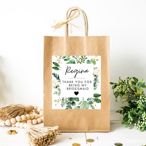 May include: A brown paper gift bag with a white tag that says "Regina Thank you for being my bridesmaid" with a small black heart. The tag has a floral design with white flowers and green leaves.
