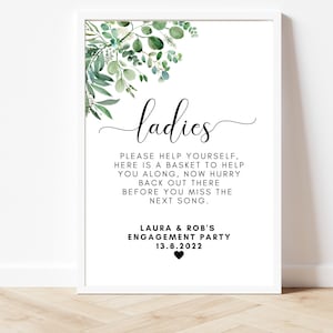Puede incluir: Un cartel blanco con follaje verde en la parte superior. El cartel dice "ladies" en una fuente cursiva. El texto debajo dice "Por favor, s&iacute;rvete, aqu&iacute; hay una cesta para ayudarte, date prisa y vuelve all&iacute; antes de que te pierdas la pr&oacute;xima canci&oacute;n." El cartel tambi&eacute;n incluye el texto "Laura & Rob's Engagement Party 13.8.2022".