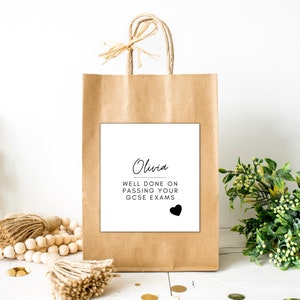 Puede incluir: Una bolsa de regalo de papel marr&oacute;n con una etiqueta cuadrada blanca. La etiqueta dice "Olivia, Bien hecho al aprobar tus ex&aacute;menes GCSE" con un coraz&oacute;n negro. La bolsa tiene un lazo de rafia y asas.