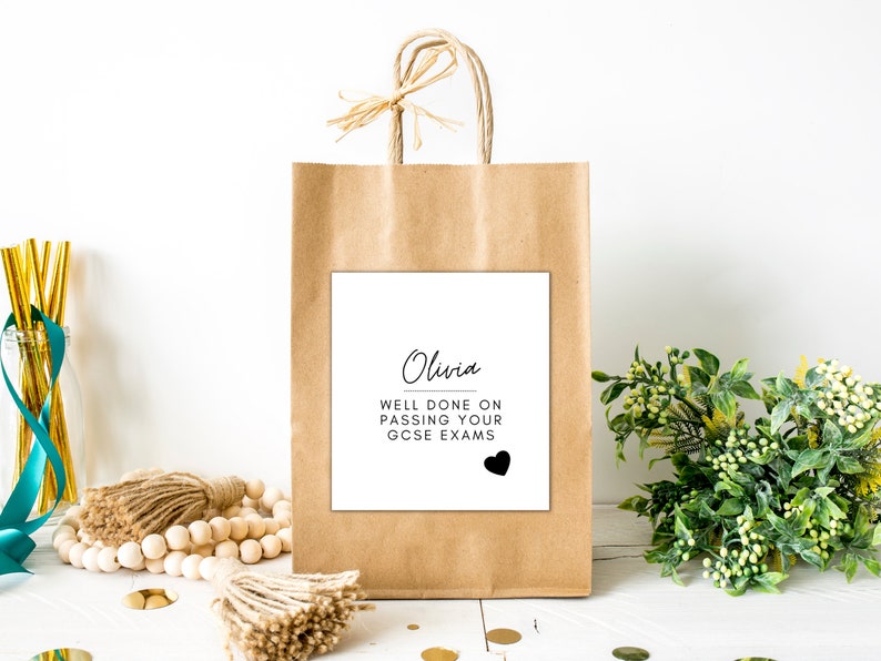 Puede incluir: Una bolsa de regalo de papel marr&oacute;n con una etiqueta cuadrada blanca. La etiqueta dice "Olivia, Bien hecho al aprobar tus ex&aacute;menes GCSE" con un coraz&oacute;n negro. La bolsa tiene un lazo de rafia y asas.