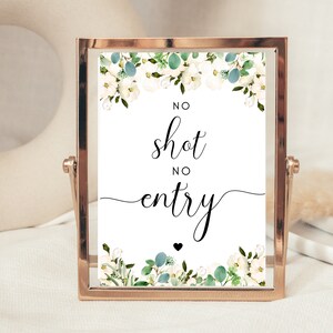 Puede incluir: Un letrero enmarcado con el texto "NO shot NO entry" en escritura elegante. El letrero est&aacute; decorado con flores blancas y follaje verde. El marco es de color oro met&aacute;lico, y el letrero se muestra sobre una superficie blanca.