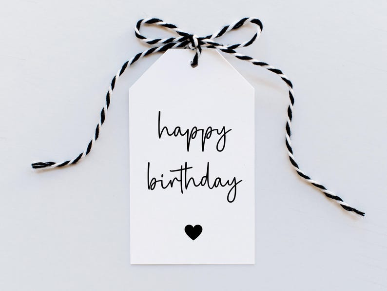 Puede incluir: Etiqueta de regalo blanca con una cuerda negra atada en un lazo. La etiqueta tiene el texto "feliz cumplea&ntilde;os" escrito en cursiva negra y un peque&ntilde;o coraz&oacute;n negro.