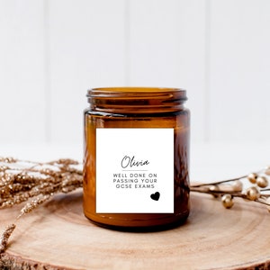 Puede incluir: Una vela en tarro de cristal marr&oacute;n con una etiqueta blanca que dice "Olivia, Bien hecho al aprobar tus ex&aacute;menes GCSE" con un coraz&oacute;n negro. La vela est&aacute; sobre una rodaja de madera con elementos decorativos dorados y marrones.