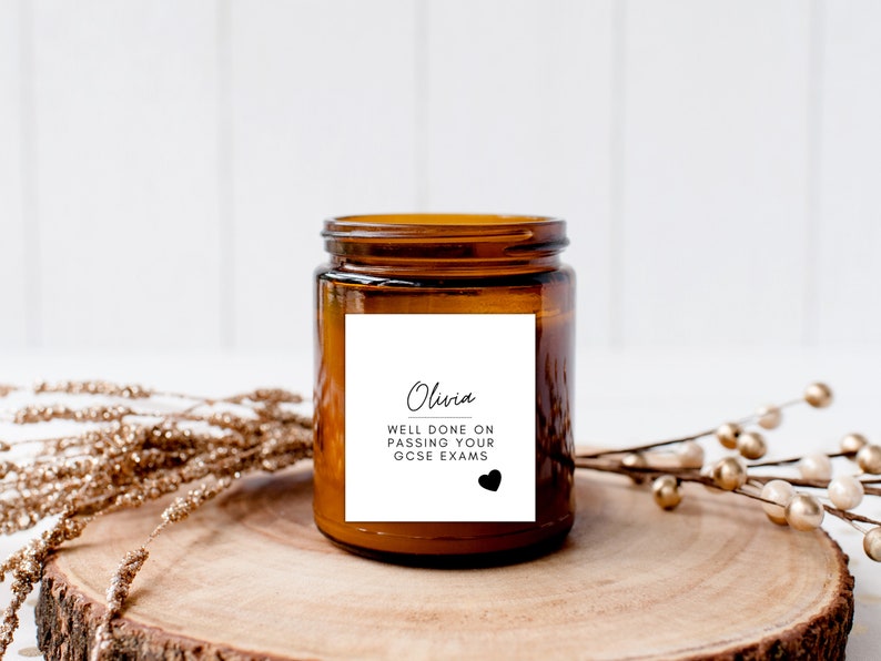 Puede incluir: Una vela en tarro de cristal marr&oacute;n con una etiqueta blanca que dice "Olivia, Bien hecho al aprobar tus ex&aacute;menes GCSE" con un coraz&oacute;n negro. La vela est&aacute; sobre una rodaja de madera con elementos decorativos dorados y marrones.