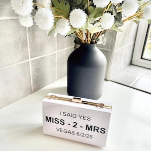 Peut inclure: Une pochette blanche avec une fermeture dorée et le texte "I said yes Miss - 2 - Mrs Vegas 6/25" imprimé sur le devant.