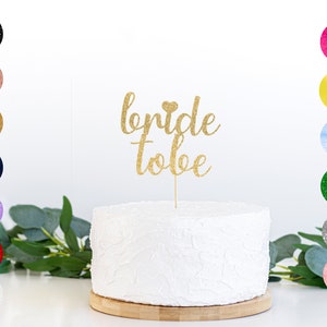May include: Gold glitter cake topper with the text "bride to be" and a small heart. The topper is on a white cake with a textured surface.