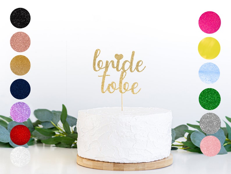 May include: Gold glitter cake topper with the text "bride to be" and a small heart. The topper is on a white cake with a textured surface.