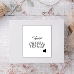 Puede incluir: Una impresi&oacute;n enmarcada en blanco con el nombre "Olivia" y el mensaje "WELL DONE ON PASSING YOUR GCSE EXAMS" en escritura negra. Un coraz&oacute;n negro en la parte inferior. La impresi&oacute;n est&aacute; sobre una superficie blanca, con una caja de regalo y cuerda en el fondo.