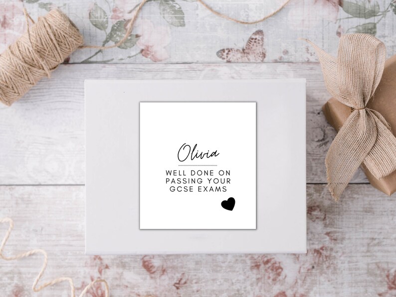 Puede incluir: Una impresi&oacute;n enmarcada en blanco con el nombre "Olivia" y el mensaje "WELL DONE ON PASSING YOUR GCSE EXAMS" en escritura negra. Un coraz&oacute;n negro en la parte inferior. La impresi&oacute;n est&aacute; sobre una superficie blanca, con una caja de regalo y cuerda en el fondo.
