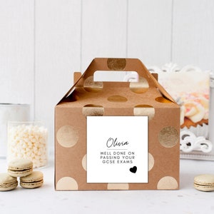 Puede incluir: Una caja de regalo marr&oacute;n con lunares dorados y un asa. Una etiqueta cuadrada blanca en la parte delantera dice "Olivia, Bien hecho al aprobar tus ex&aacute;menes GCSE" con un coraz&oacute;n negro. Macarons y objetos decorativos en el fondo.
