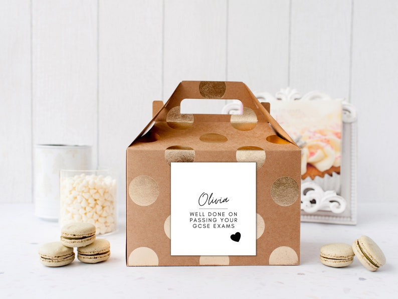 Puede incluir: Una caja de regalo marr&oacute;n con lunares dorados y un asa. Una etiqueta cuadrada blanca en la parte delantera dice "Olivia, Bien hecho al aprobar tus ex&aacute;menes GCSE" con un coraz&oacute;n negro. Macarons y objetos decorativos en el fondo.