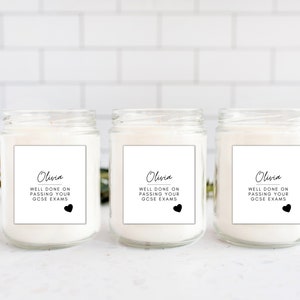 Puede incluir: Tres velas en tarro de cristal con cera blanca. Cada vela tiene una etiqueta blanca con el nombre "Olivia", el texto "WELL DONE ON PASSING YOUR GCSE EXAMS" y un coraz&oacute;n negro. Los tarros est&aacute;n sobre una superficie blanca.