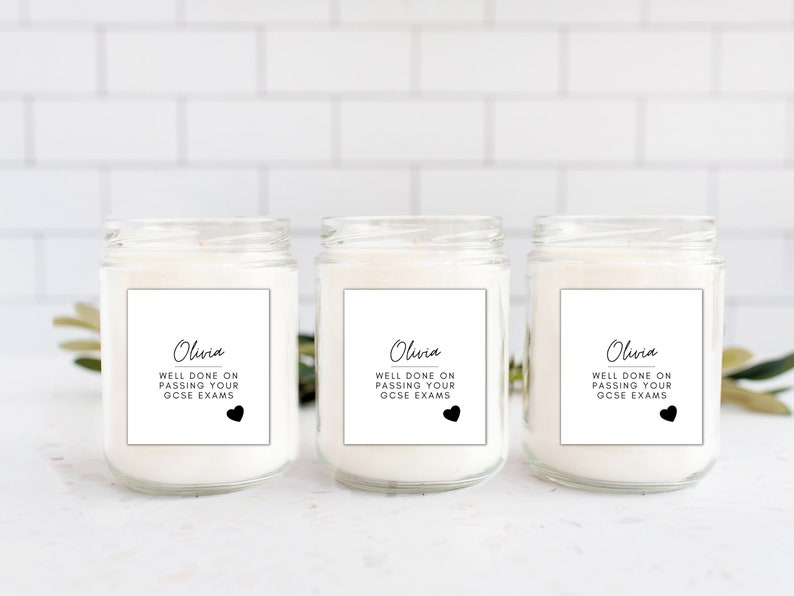 Puede incluir: Tres velas en tarro de cristal con cera blanca. Cada vela tiene una etiqueta blanca con el nombre "Olivia", el texto "WELL DONE ON PASSING YOUR GCSE EXAMS" y un coraz&oacute;n negro. Los tarros est&aacute;n sobre una superficie blanca.