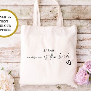 May include: White canvas tote bag with the text "SARAH cousin of the bride" and a small heart. The bag has a gold circle with the text "OVER 10 TEXT COLOUR OPTIONS".