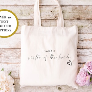 May include: White canvas tote bag with black text that reads "SARAH sister of the bride" with a small heart.