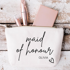 以下が含まれることがあります： 「maid of honour Olivia」と小さなハートが書かれた黒い文字の白いキャンバス製メイクアップバッグ。