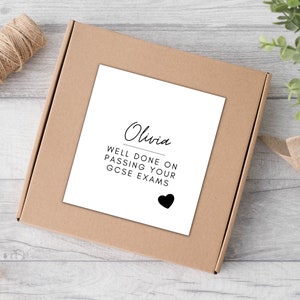 Puede incluir: Una caja de cart&oacute;n marr&oacute;n con una tarjeta blanca. La tarjeta dice "Olivia, BIEN HECHO AL APROBAR TUS EX&Aacute;MENES GCSE" con un coraz&oacute;n negro. Tambi&eacute;n se ven hilo y vegetaci&oacute;n.