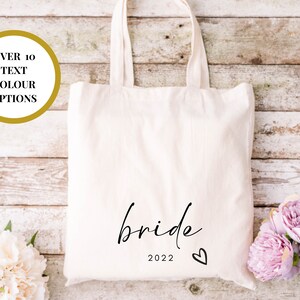 May include: White canvas tote bag with the word "bride" in black script and the year "2022" below it. A small heart is to the right of the year.
