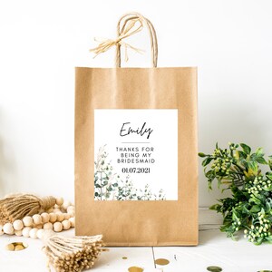 May include: A brown paper gift bag with a white label that says "Emily Thanks for being my bridesmaid 01.07.2021" with a green floral design.